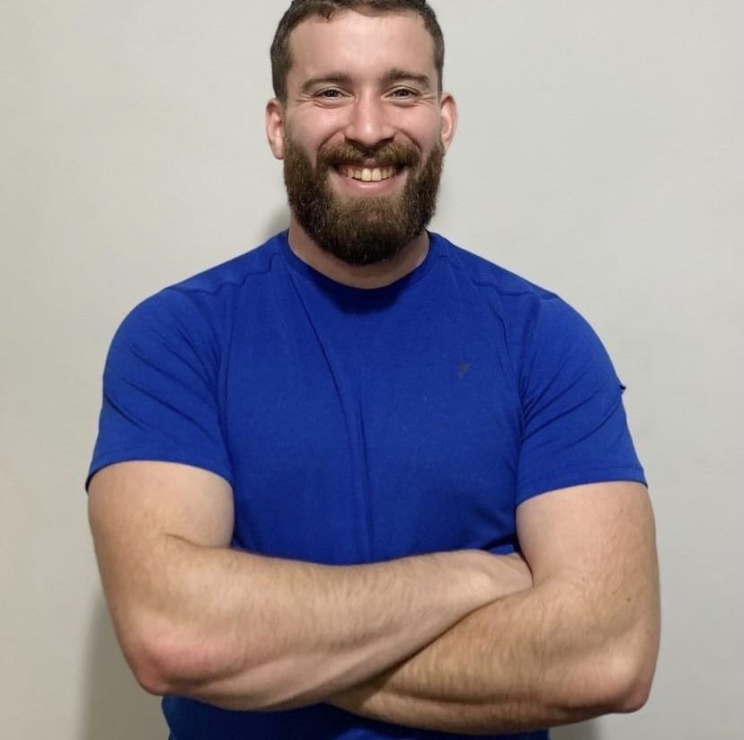 The image shows a man standing against a neutral-colored background. He has a full beard and short hair. He is smiling and has his arms crossed over his chest. He is wearing a fitted blue t-shirt. The lighting in the room is soft, and the photo appears to be candid.